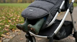 A detailed photorealistic close-up on a damp autumnal day in a British park, showing muddy toddler wellington boots sticking out of a practical, unzipped opening at the foot of a charcoal grey quilted pram footmuff.