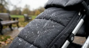 A close-up photorealistic illustration of a charcoal grey quilted footmuff fabric during a rainy UK autumn day, with distinct water droplets beading and running off the surface.