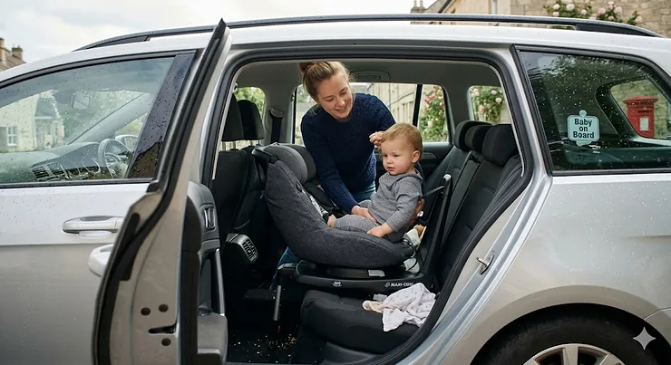 A 360 rotating car seat turned towards the open door of a silver car, showing the easy-access swivel base for loading a child in a typical UK village setting. 360 rotating car seat