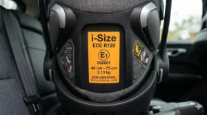 A highly detailed close-up photograph of the orange ECE R129 i-Size approval label affixed to the plastic shell of a car seat, clearly displaying the required technical safety information, height, and weight limits for use in the UK.