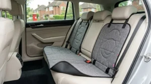 Full rear bench car seat protector mat in durable grey fabric laid flat across cream leather upholstery showing alignment slits for ISOFIX anchors and seatbelt receivers.